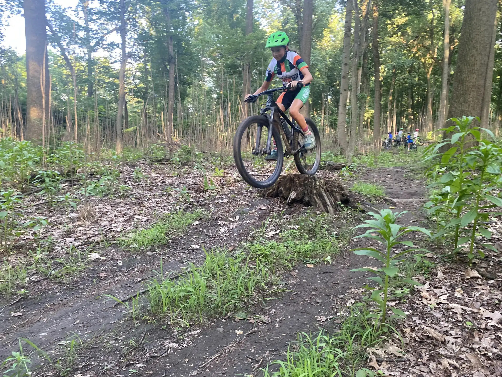 A helmeted teammate rides over a small wooded trail feature while other riders wait among the trees.